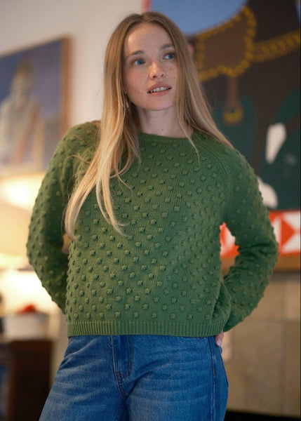 A woman with long blonde hair wearing a textured olive green knit sweater and blue jeans, standing in a warmly lit indoor setting with framed artwork in the background.