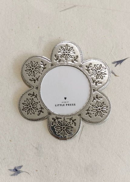 A silver-toned metal flower-shaped photo frame with embossed floral patterns, displayed on a textured cream fabric background. The center is blank, and the brand name 'LITTLE PRESS' is printed on the front.