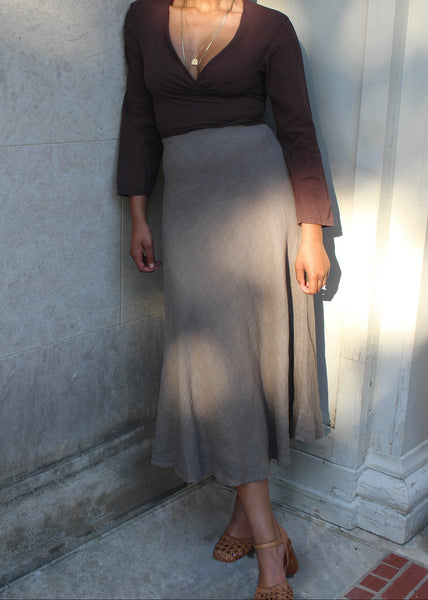 Woman wearing a brown top and gray skirt standing against a light-colored wall.