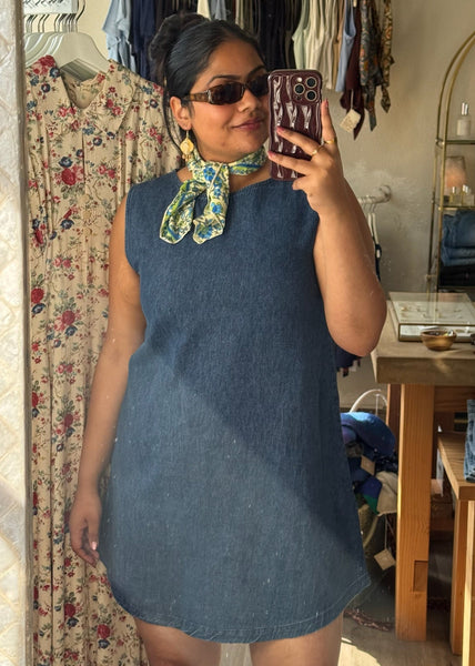 Woman taking a mirror selfie wearing a blue dress in a room with clothing racks and furniture.