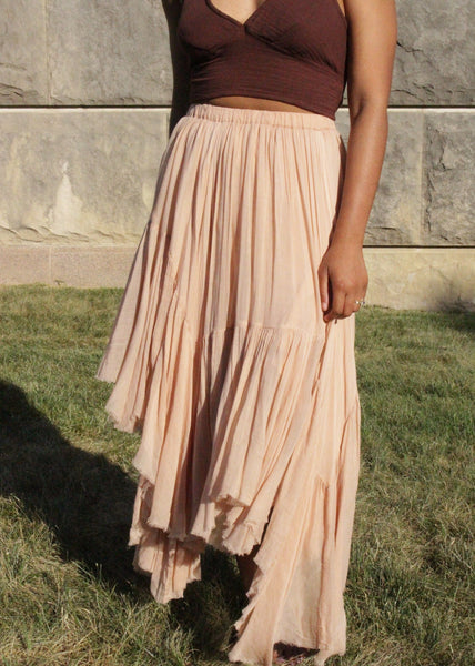 Person wearing a brown top and beige pleated skirt standing on grass with a stone wall background