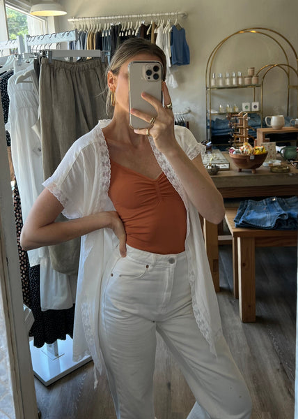 Person taking a mirror selfie wearing a white cardigan, orange top, and white pants in a clothing store.