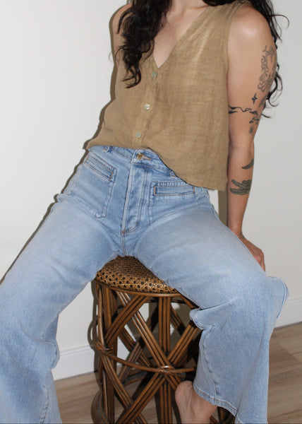 Person wearing a beige sleeveless top and blue jeans sitting on a wooden stool.