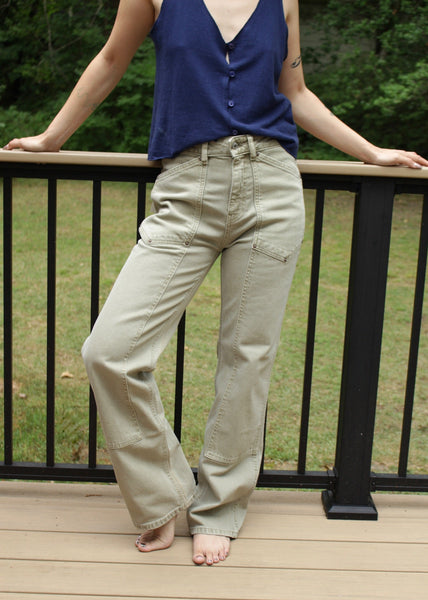 Person wearing a blue top and beige pants standing on a wooden deck with greenery in the background