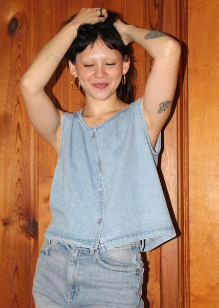 Person wearing a denim sleeveless top and jeans against a wooden wall.