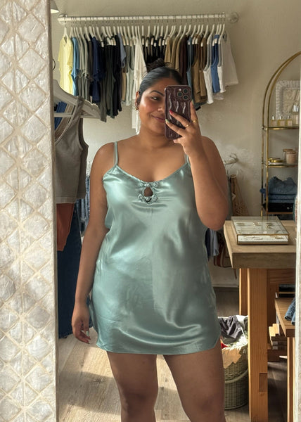 Person wearing a light blue satin nightgown taking a mirror selfie in a room with clothes on hangers.
