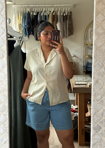 Woman taking a mirror selfie wearing a white shirt and blue shorts in a room with clothing racks.