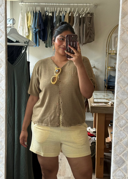 Woman taking a mirror selfie wearing a beige cardigan and light-colored shorts in a room with clothing racks.
