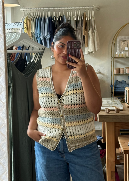 A woman takes a mirror selfie wearing a handmade crochet vest with pastel stripes of blue, yellow, pink, and beige, paired with blue jeans in what appears to be a boutique setting.