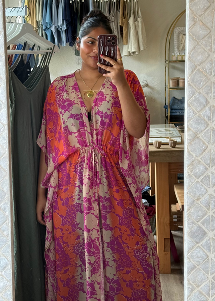 A woman takes a mirror selfie in a boutique showing a loose, long-sleeve kaftan dress with a vibrant magenta, orange, and beige floral pattern.