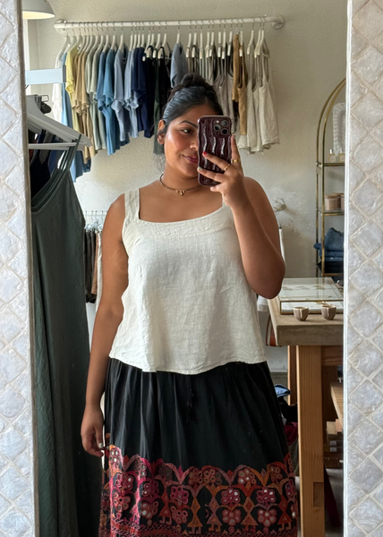 A woman takes a mirror selfie in a boutique while wearing a white sleeveless crop top and a long black skirt with a colorful red, orange, and gold bohemian patterned hem.