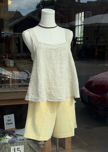 A two-piece linen set displayed on a mannequin in a storefront window. The set includes a cream-colored sleeveless top and light yellow shorts.