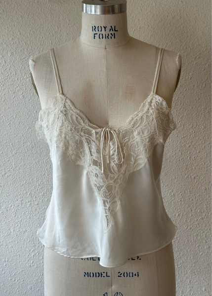 An ivory satin camisole top with lace cups and a front tie, displayed on a vintage fabric dress form mannequin against a textured wall.