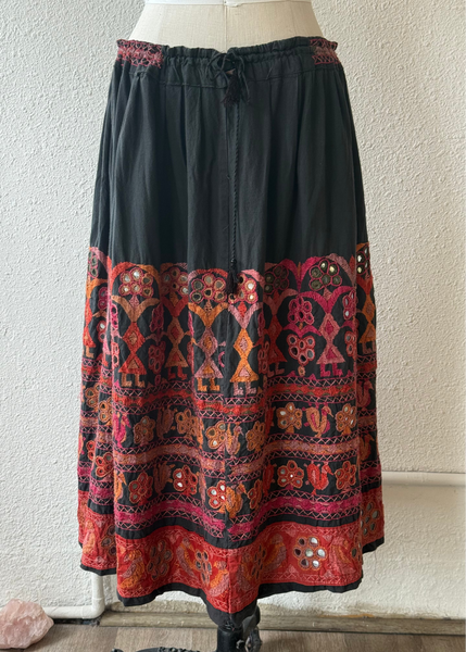 A black midi skirt with a highly detailed, colorful bohemian-style border print in red, orange, and pink, displayed on a mannequin against a textured off-white wall.
