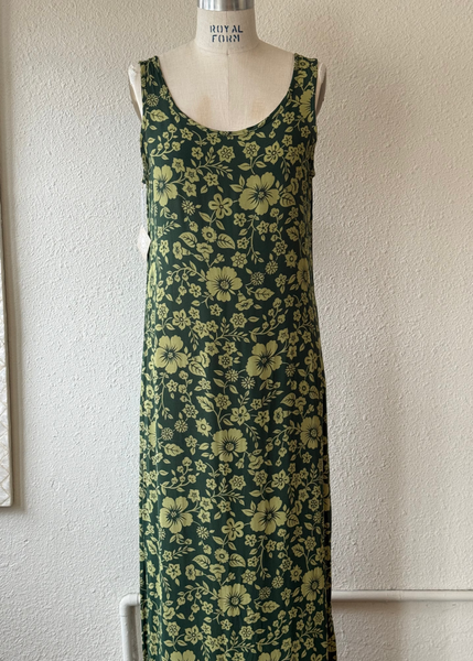 A sleeveless scoop-neck maxi dress with a green and olive yellow floral print, displayed on a mannequin torso against a textured off-white wall.