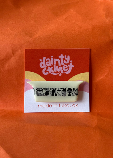 A small enamel pin mounted on a branded display card. The pin is cream-colored with black illustrations of various fruits and vegetables, and the card reads 'dainty comet' and 'made in tulsa, ok'.
