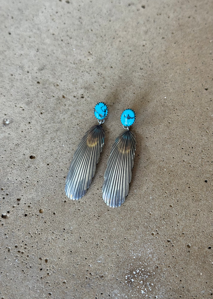 A pair of silver-toned dangle earrings with oval turquoise blue stone tops and ribbed feather-like drops, photographed on a textured concrete surface.