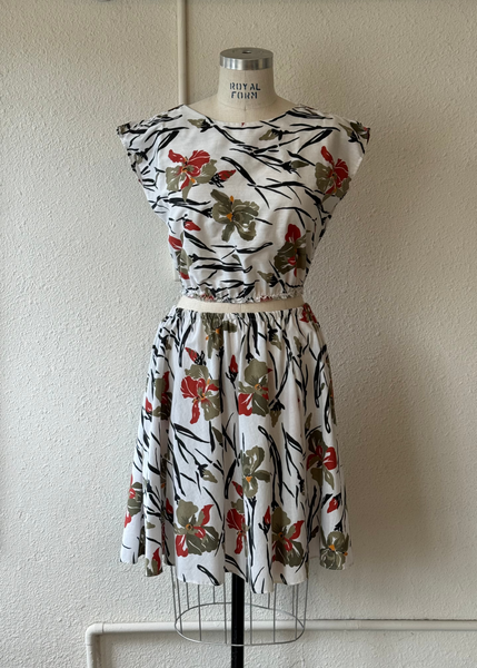 A sleeveless two-piece dress set displayed on a dress form mannequin against a textured off-white wall. The set features a short-sleeve top and matching flared skirt with a white base and a botanical print of red flowers, olive green leaves, and black branch details.