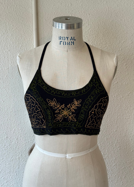 A black halter-style bralette with gold and green embroidered floral patterns, displayed on a cream-colored dress form against a textured off-white wall.