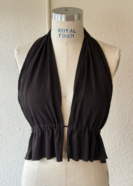 A black halter-style top displayed on a cream-colored mannequin dress form against a textured off-white wall. The top features a gathered, ruffled hem and a deep V-neckline.