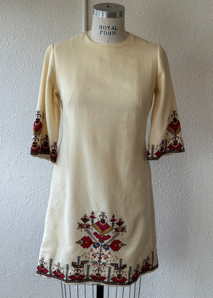 An off-white/cream short dress with red, blue, and black embroidered patterns on the cuffs and hem, displayed on a mannequin torso against a textured white wall.