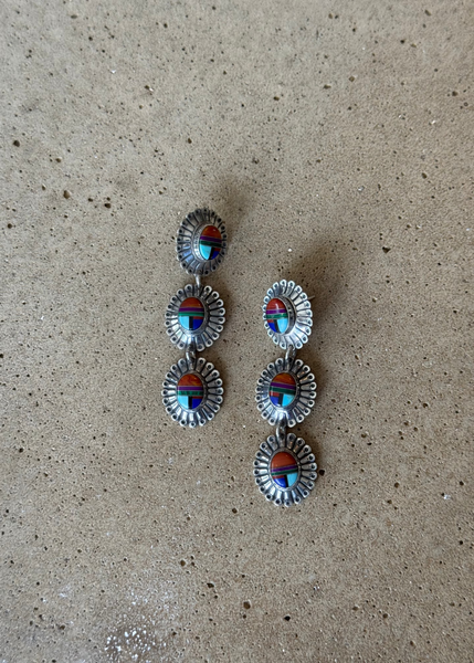 A pair of silver-toned drop earrings with three oval stacked charms, each featuring a colorful inlay of turquoise, red, and blue stones, displayed on a textured beige surface.