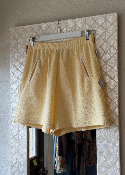 A pair of light yellow textured lounge shorts with side pockets, hanging from a white hanger on a patterned tile wall.