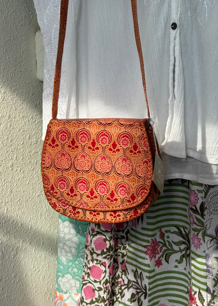 A small shoulder bag with a brown leather strap and a vibrant orange, red, and pink patterned fabric body, shown hanging against a white button-up shirt and a floral skirt.