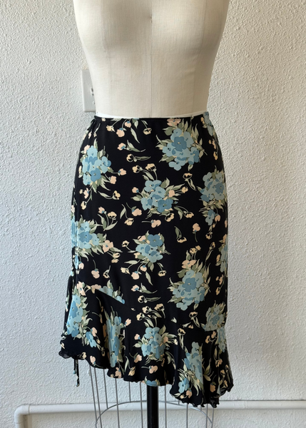 A black skirt with a light blue and beige floral pattern, displayed on a cream-colored dress form mannequin against a textured white wall.