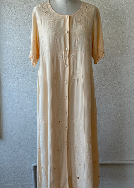A cream-colored cotton nightgown with floral embroidery and button-front closure, displayed on a mannequin torso against a textured white wall.