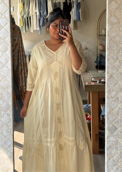 A woman takes a mirror selfie in a boutique wearing a cream-colored, tiered cotton maxi dress with embroidered details. The background shows a clothing rack filled with various garments.