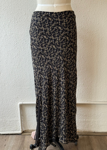 A long black maxi skirt with a beige floral print, displayed on a dress form mannequin against a textured off-white wall.