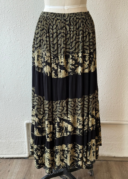 A long tiered maxi skirt displayed on a dress form mannequin, featuring a black and beige bohemian-style floral and abstract pattern with solid black horizontal bands.