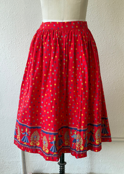 A red A-line skirt with a colorful polka dot pattern and a decorative embroidered border, displayed on a cream dress form mannequin against a textured off-white wall.