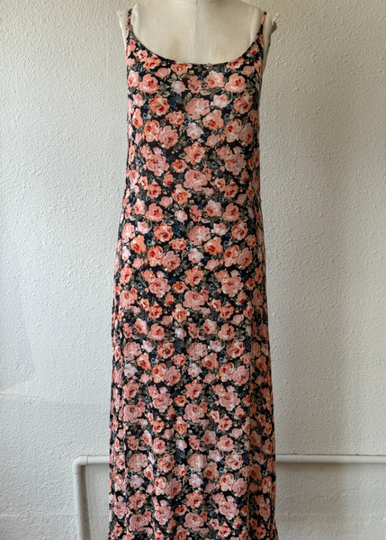 A sleeveless floral maxi dress displayed on a dress form against a textured off-white wall. The dress features a dense, all-over floral pattern in shades of pink, peach, and white on a very dark, near-black background.