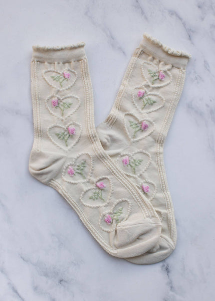 A pair of cream-colored casual socks with embossed heart shapes and adorned with colorful roses, displayed against a marble background.