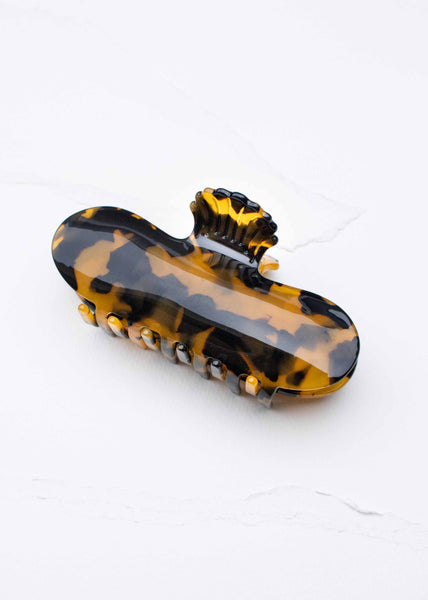 A tortoiseshell hair claw clip with a glossy black and amber-brown pattern, shown on a light off-white textured background.